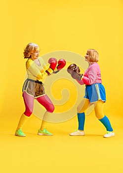 Keeping strong and fit. Two elderly sportive woman in colorful uniform training, boxing, posing against yellow studio