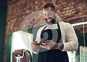 Keeping up with the new hair trends. a handsome young barber standing alone in his salon and using his cellphone.