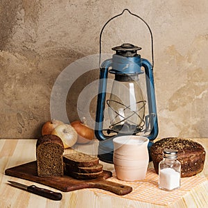 Kerosene lantern, cutting board, clay pot with milk, rye bread, salt and salt salt, and onion on a wooden table