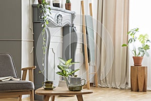 Kettle on wooden table next to grey armchair in retro living room interior with plants. Real photo