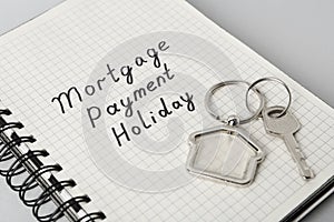 Key with trinket in shape of house and phrase Mortgage payment holiday written on notebook against light grey background, closeup