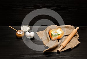 Khachapuri with eggs on sackcloth, salt, flour, eggs and parsley on the black table, side view