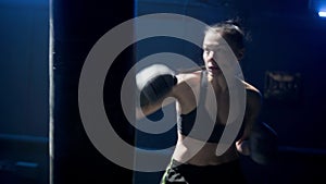 Kickboxing training in dark gym with blue light. Young woman fighter in black uniform, boxing gloves and leg protection