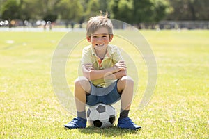 Kid 7 or 8 years old enjoying happy playing football soccer at grass city park field posing smiling proud sitting on the ball in