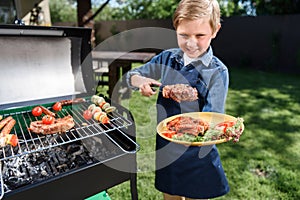 Kid boy in apron preparing tasty stakes on barbecue grill outdoors