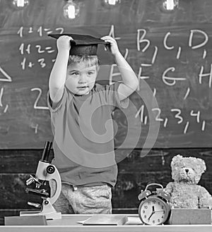 Kid boy in graduate cap ready to go to school, chalkboard on background. Child, pupil on smiling face near microscope