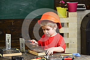 Kid boy in orange hard hat or helmet, study room background. Boy play as builder or repairer, work with tools. Child