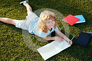 Kid boy with pencil writing on notebook lying on grass. Cute little child in casual clothes reading a book and smiling