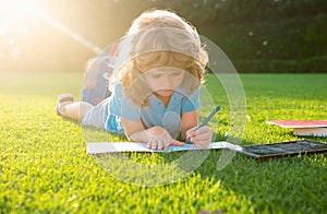 Kid boy reading a book lying on grass writing notes in copybook. Cute little child in casual clothes reading a book in