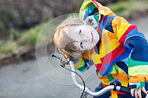 Kid boy in safety helmet and colorful raincoat riding bike, outd