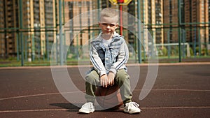 Kid boy is sitting on basketball ball on court in residental district in city.