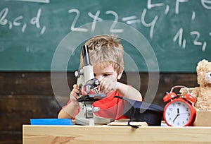 Kid boy work with microscope in classroom, chalkboard on background. Child on busy face near clock and teddy bear