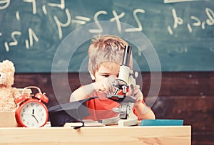 Kid boy work with microscope in classroom, chalkboard on background. Child on busy face near clock and teddy bear