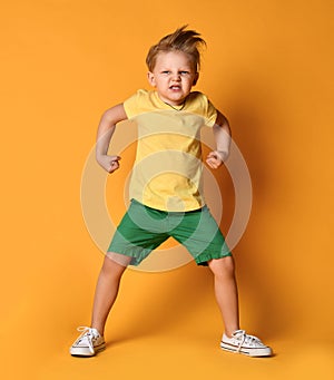 Kid boy in yellow t-shirt, green short and white sneakers is in a pose of angry monster demonstrating power and hisses with rage