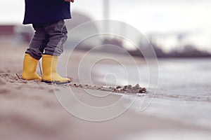 A kid in bright yellow rubber boots is playing at the surf zone of a sandy sea shore in front of a nuclear power plant