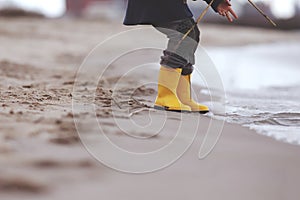 A kid in bright yellow rubber boots is playing at the surf zone of a sandy sea shore