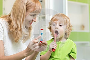 Kid brushing teeth near mirror in bathroom. His mother monitoring accuracy and time of cleaning action with hourglass.