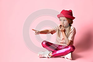Kid in casual outfit is looking surprised, pointing by forefinger at something, sitting on floor with crossed legs against pink