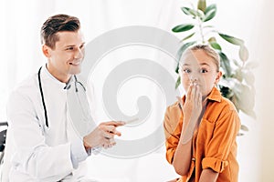 Kid covering mouth with hand and smiling pediatrist holding medical spatula