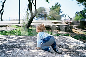 Kid crawls along the stone platform against the backdrop of green trees. Back view