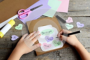 Kid doing a greeting card. Kid holds a black marker in hand and writes wishes. Paper greeting card with jar, hearts and wishes