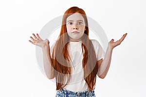 Kid dont know. Cute redhead little girl shrugging and raise hands up, look clueless and confused at camera, have no idea
