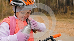 Kid drink water from an aluminum flask. One caucasian children rides bike road in autumn park. Little girl riding black orange mtb