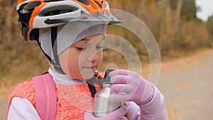 Kid drink water from an aluminum flask. One caucasian children rides bike road in autumn park. Little girl riding black