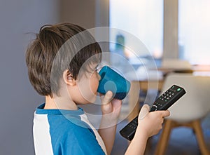 Kid drinking soda drink in plastic glass and holding remote control, Asian Young boy having fun drinking soft drink and watchung