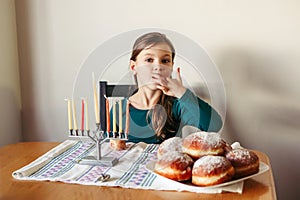 Kid eating sufganiyot donuts, sweet festive food. Girl with menorah for traditional winter Jewish Hanukkah holiday. Child