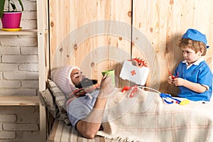 Kid and father playing hospital. Patient with flu lying in bed under woolen blanket. Doctor visiting sick bearded man at