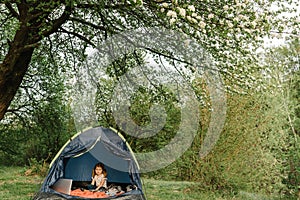 Kid girl in a campaign in a tent. Family summer vacation in nature. Children tourism. Child using laptop in the tent at the