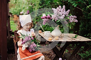 Kid girl at garden tea party in spring day with bouquet of lilacs syringa