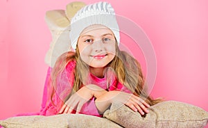 Kid girl knitted hat. Winter accessory concept. Girl long hair dream pink background. Kid dreamy lean on pillows. Winter