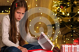 Kid girl opening Christmas gift in decorated room and gets upset