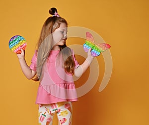 Kid girl in pink clothes shows two sensory rainbow color toys - pop it. Holds round and looks at butterfly shape one