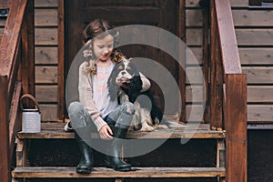 Kid girl playing with her spaniel dog, sitting on stairs at wooden log cabin