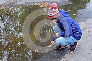 Kid girl in a warm knitted cap playing with leaves near a puddle while walking in an autumn park.