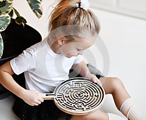 Kid girl in white t-shirt and black skirt plays educational game interestedly with wooden labyrinth