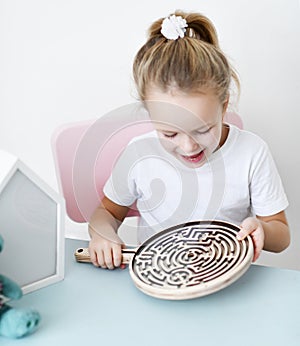 Kid girl in white t-shirt sits at the desk and plays educational game interestedly with wooden labyrinth