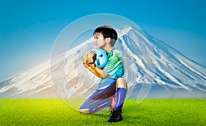 Kid goalkeeper holding the ball on a grass field with Fuji mountain background for Japanese youth soccer