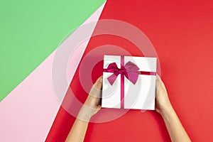Kid hands holding present gift box with ribbon on red green and pink background. Congratulations with Christmas, mother