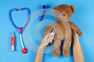 Kid hands measuring temperature with toy thermometer of teddy bear, toys medicine tools on light blue background. Top