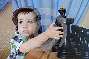 A kid in a hat with serious eyes looks to the side sitting a chess table with figures and holding a horse with hand.