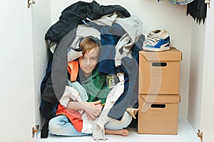 Kid hiding among messy clothes inside closet. Organization and storage of clothes at home