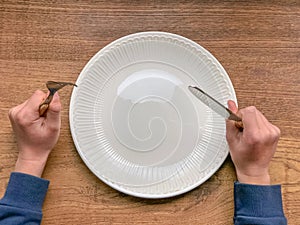 A kid is hungry. An empty white plate on a wooden table. Fork and knife. Fasting, dieting concept