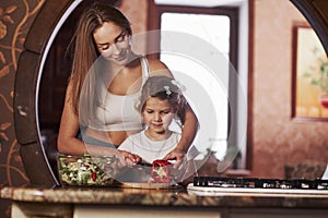 Kid learns fast. Pretty young woman standing in the modern kitchen near gas stove and teaches daughter to prepare food