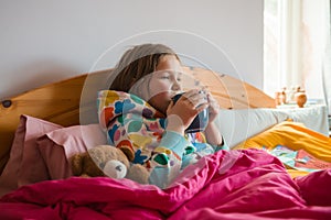 Kid lying in bed with pink blanket. Little girl with cold in scarf and blanket with hot beverage in hands near teddy