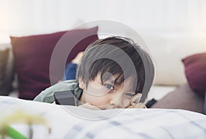 Kid lying on sofa looking at camera with thinking face, Cute boy laying down on couch holding remote, Child watching TV relaxing