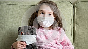 Kid makes faces and has fun at home due to COVID-19 coronavirus pandemic. Portrait of funny little girl in medical mask for